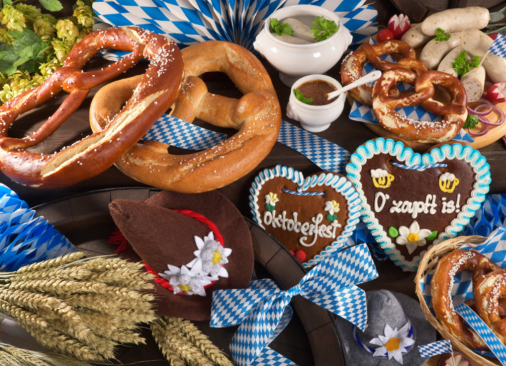Auf einem Tisch befinden sich typische bayrische Wiesnschmankerl. Bretzeln, Lebkuchenherzen, Süßer Senf auf blau-weißer Tischdecke.